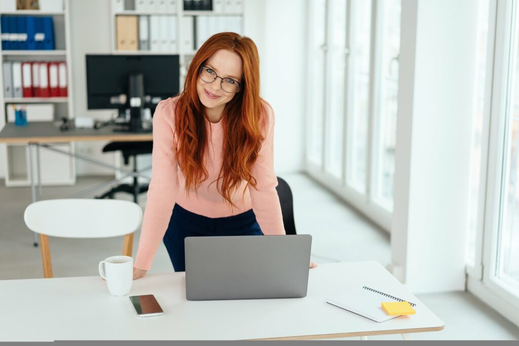Frau mit roten Haaren und einer Brille, die in einem Büro am Schreibtisch steht